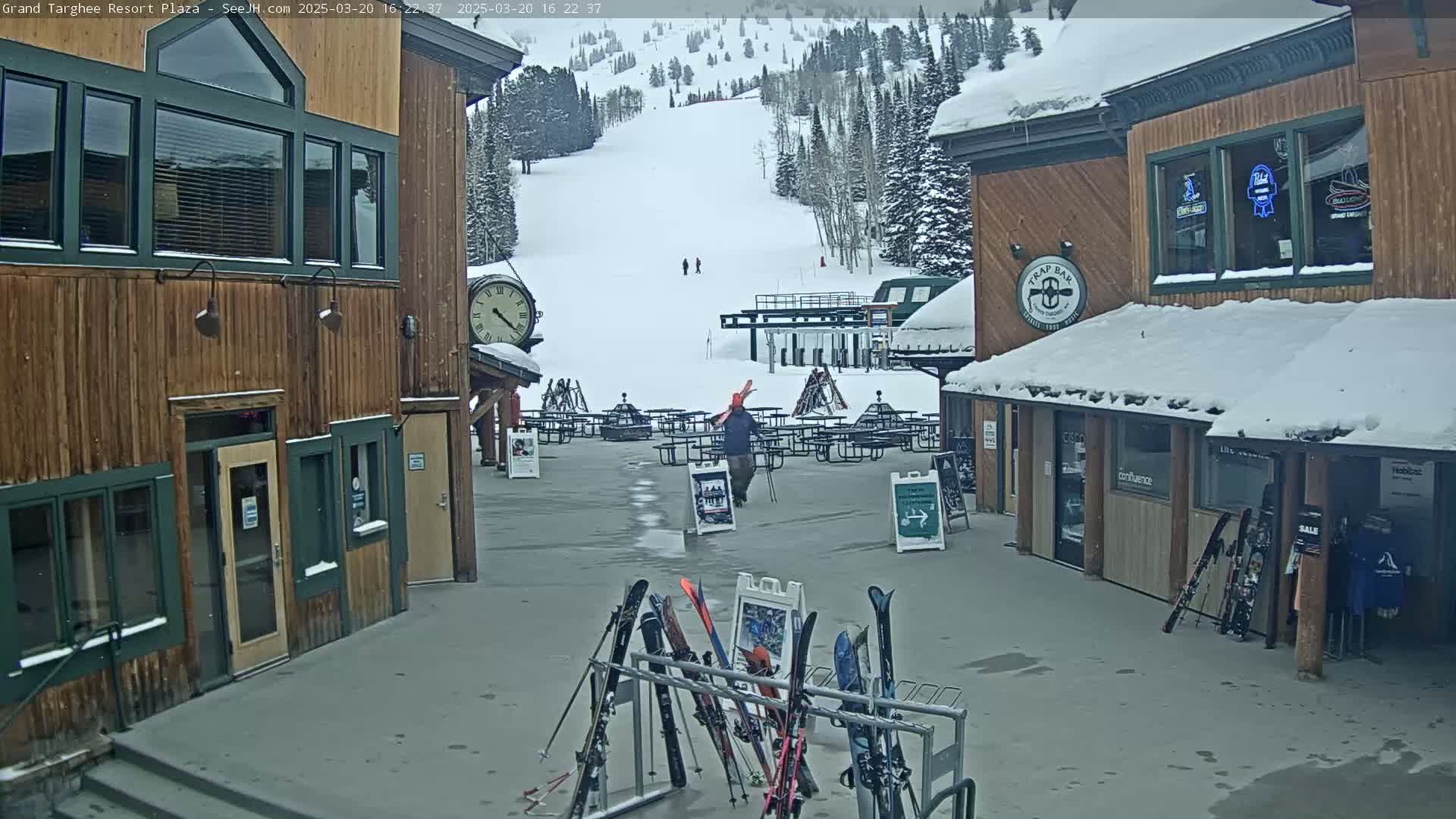 A snow-covered plaza between two wooden buildings overlooks a ski slope with several skiers visible in the distance.