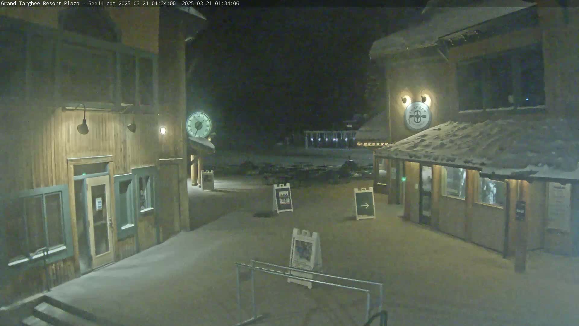 A snow-covered plaza at night, illuminated by lights on wooden buildings, with several signs and a clock visible.