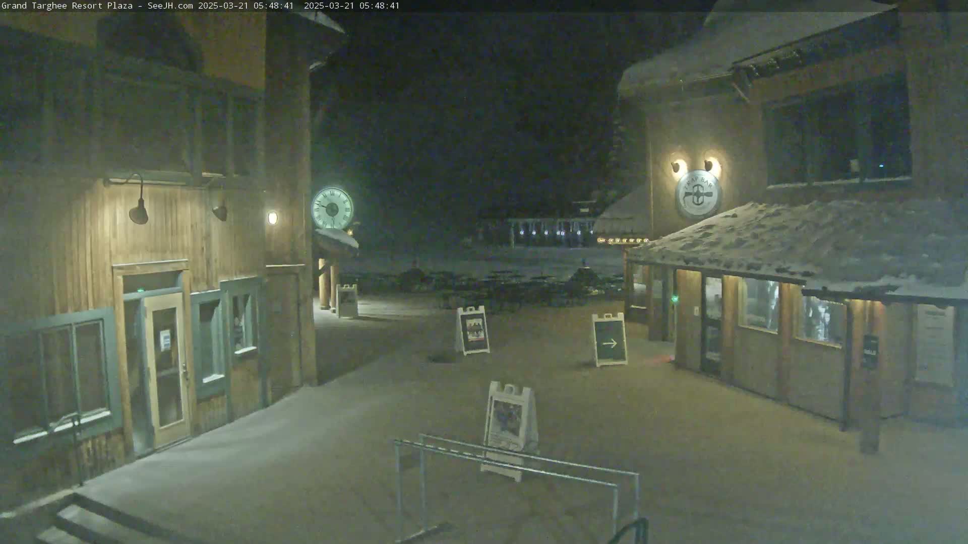 A snow-covered plaza at night, with two wooden buildings and several signs, is illuminated by dim lighting.