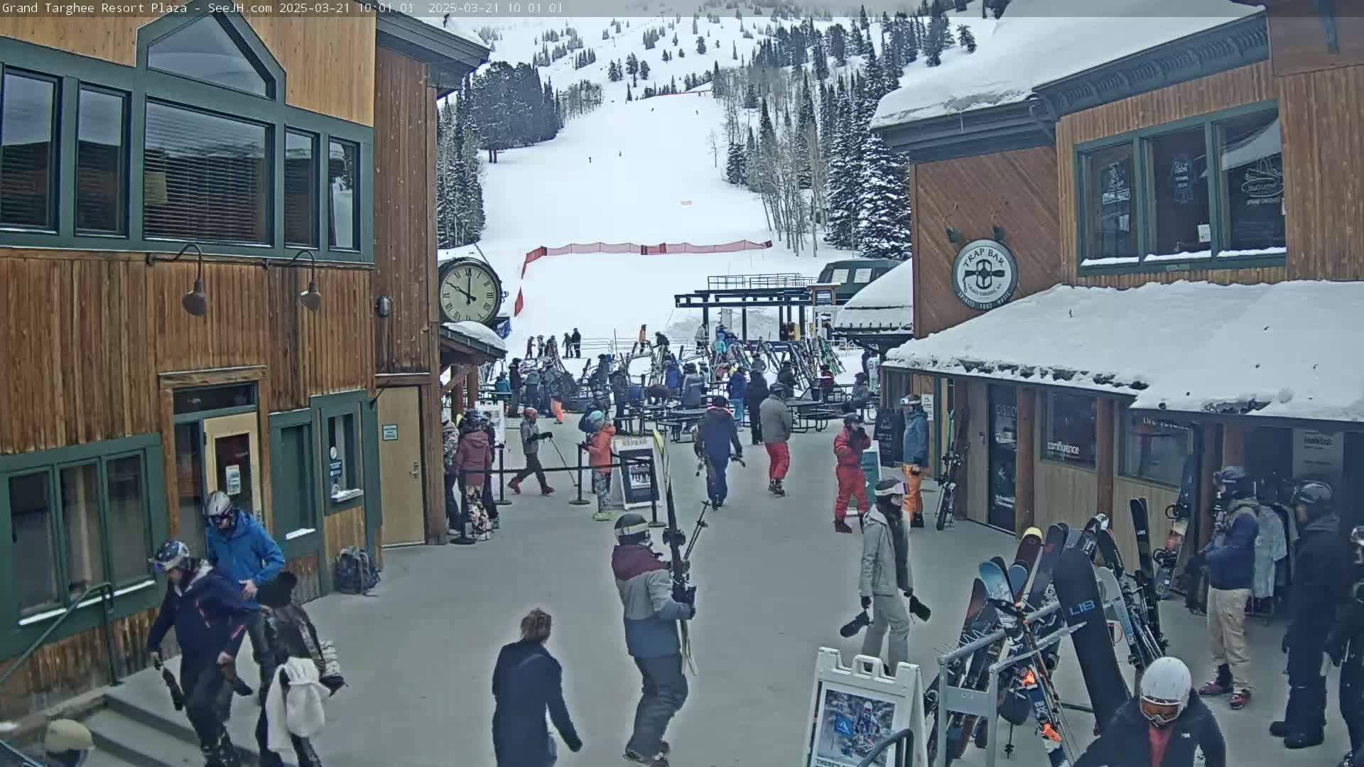 A snowy plaza at a ski resort with several buildings, people walking around, and skis leaning against a rack, with a ski slope visible in the background.