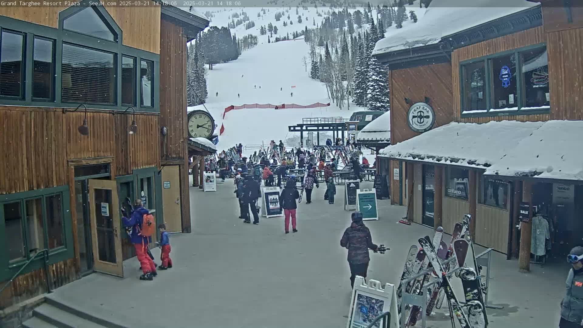 A snow-covered plaza with several wooden buildings, people, and a ski slope visible in the background under light snowfall.