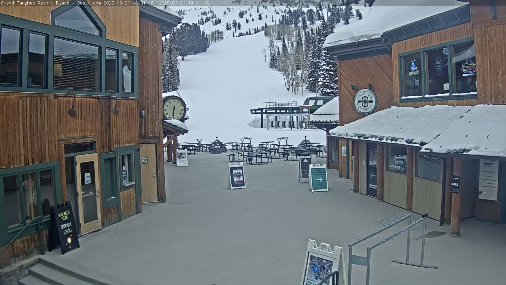 A plaza with several wooden buildings, picnic tables covered in snow, and a ski lift in the background on a snowy day.