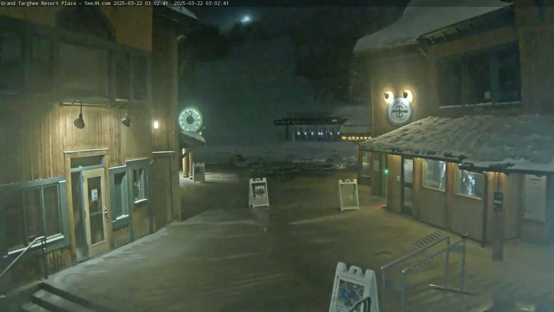 A snow-covered plaza at night, illuminated by lights on wooden buildings, shows a mostly empty square with several signs and a clock.