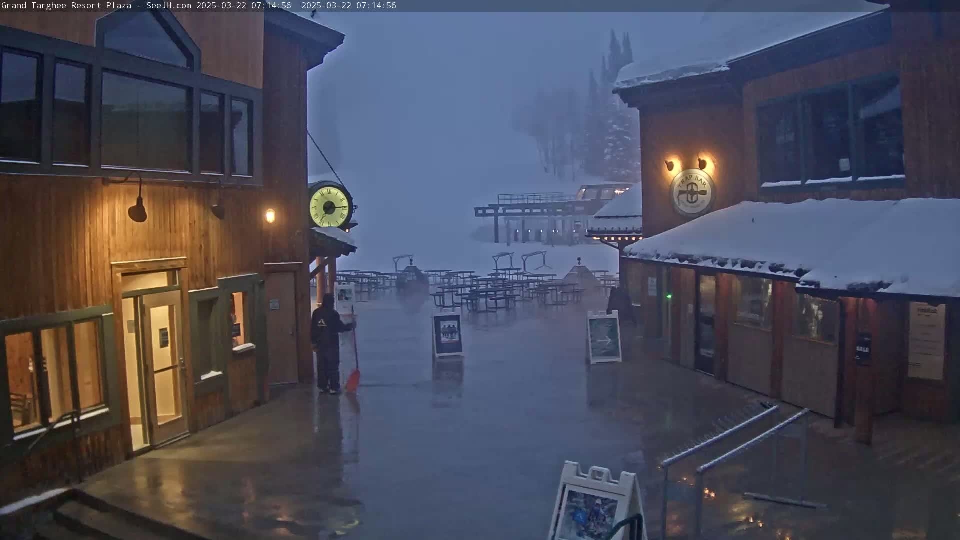 A plaza with wooden buildings, outdoor seating, and a person sweeping a wet, snow-covered surface in foggy conditions.