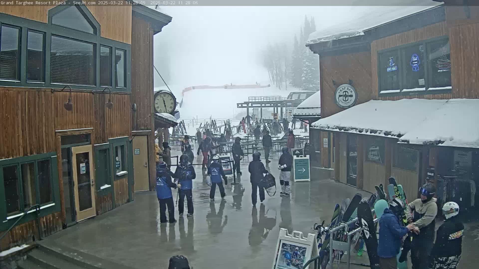 A plaza at a ski resort, during a snowy, overcast day, shows people milling about near buildings and a ski rack.