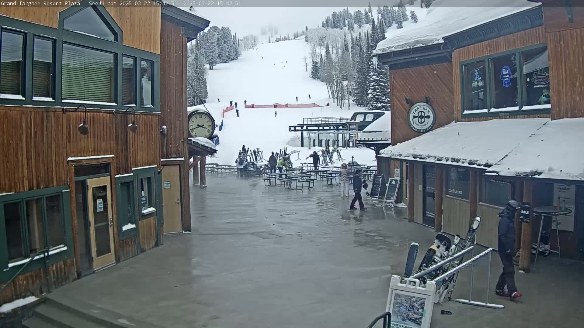 A snow-covered plaza with several buildings, outdoor seating, and skiers visible on a nearby slope under an overcast sky.