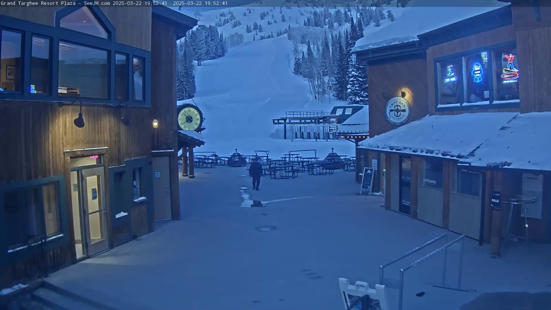 A snow-covered plaza with wooden buildings, picnic tables, and a ski slope in the background under a twilight sky.