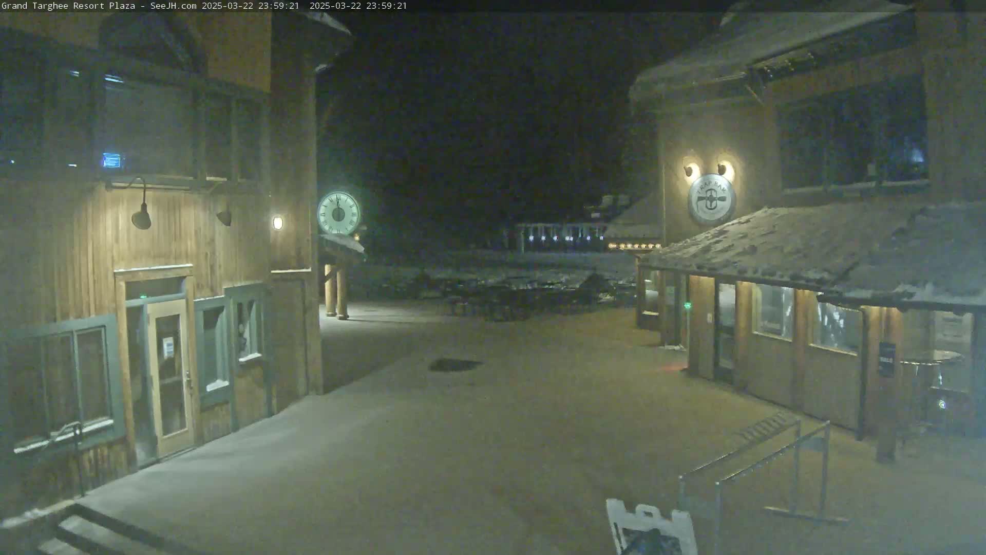 A snow-covered plaza at night is lit by outdoor lamps, showing two wooden buildings with entrances and windows.