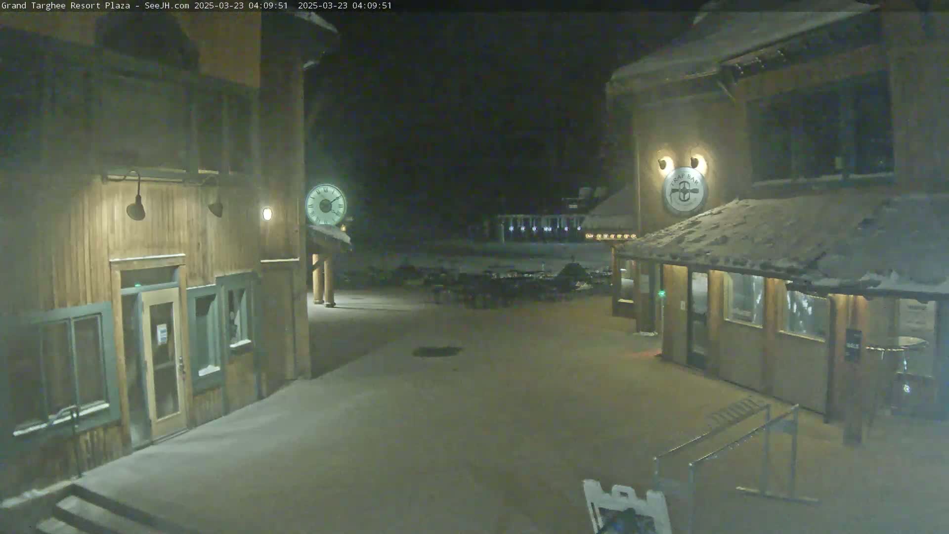 A snow-covered plaza at night is illuminated by exterior lights, with two wooden buildings flanking a sparsely populated seating area.