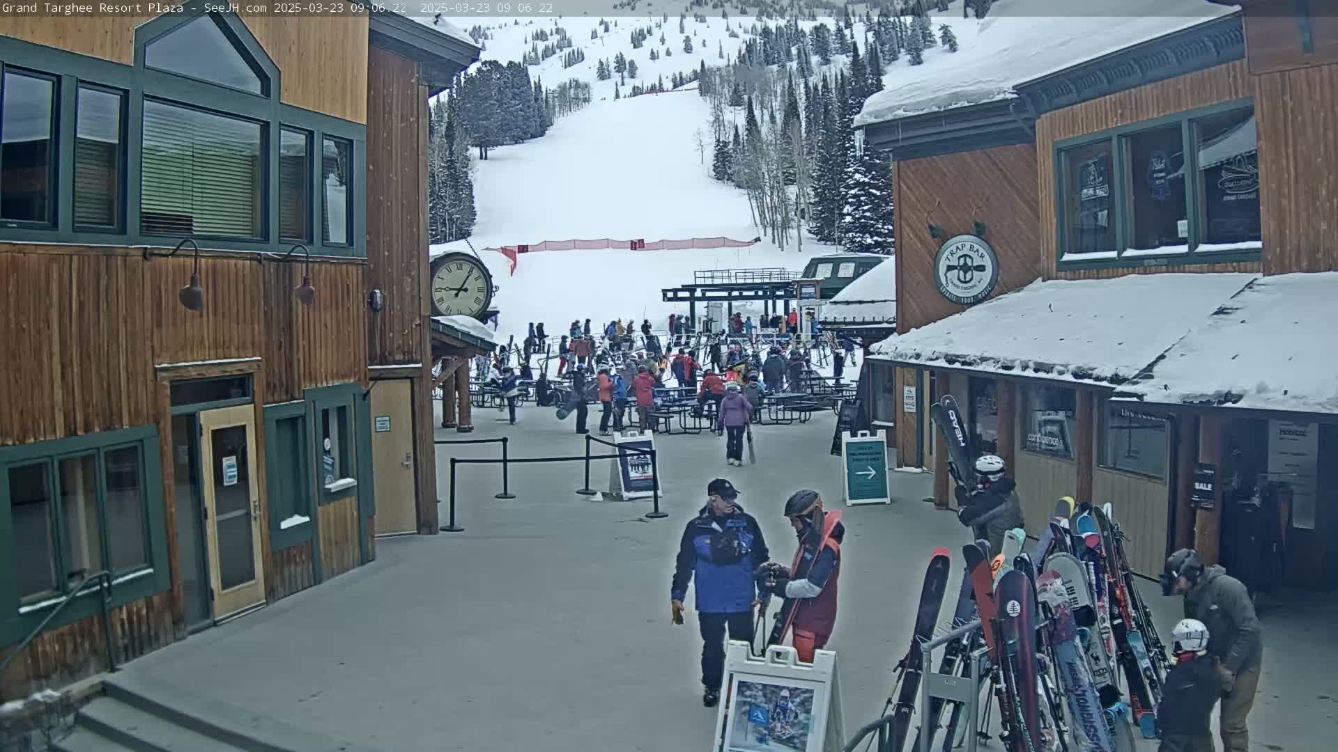 A plaza at a ski resort with several buildings, many people gathered in the area, and a snow-covered ski slope in the background under an overcast sky.