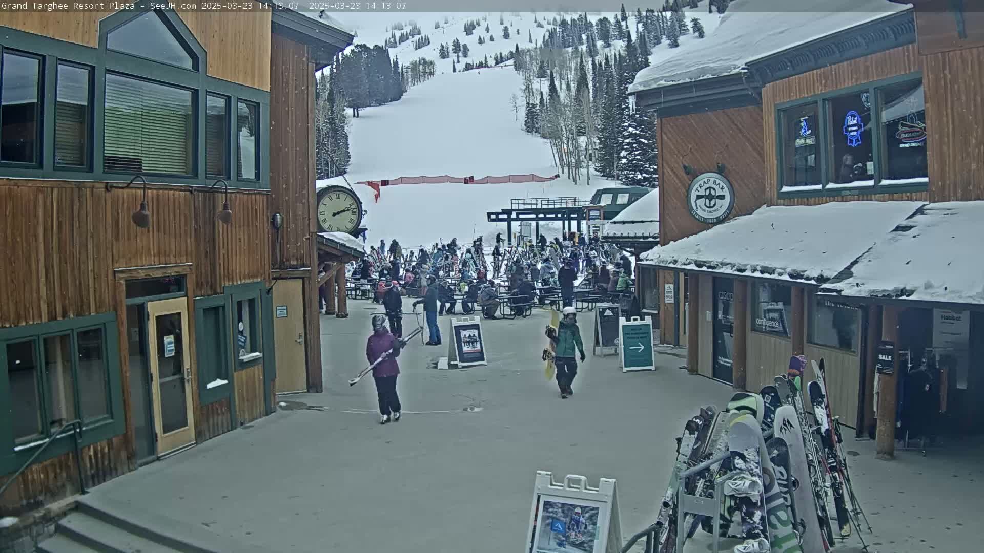 A snowy plaza scene shows several wooden buildings, a ski slope in the background, and skiers and snowboarders walking around.