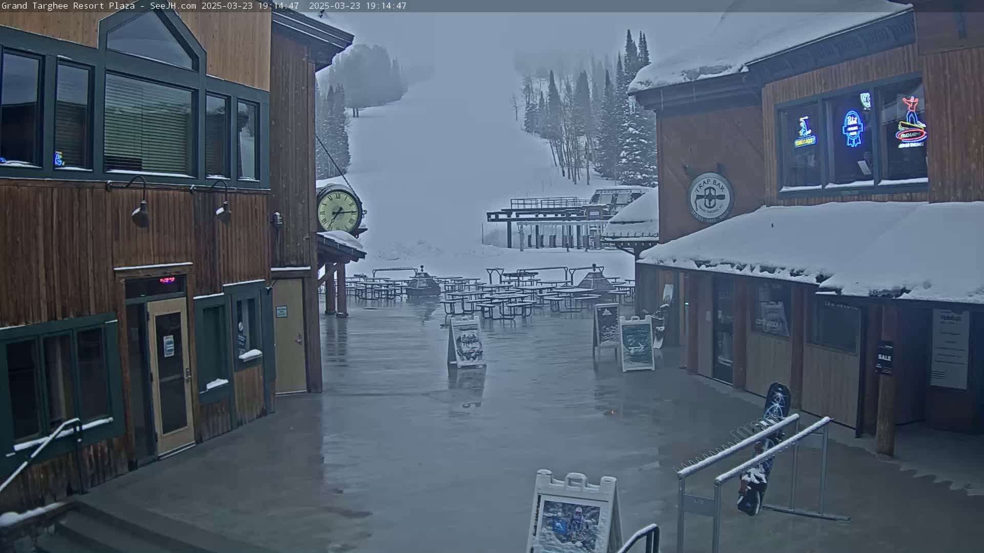 A snow-covered plaza with wooden buildings, empty picnic tables, and a ski slope visible in the background under overcast skies.