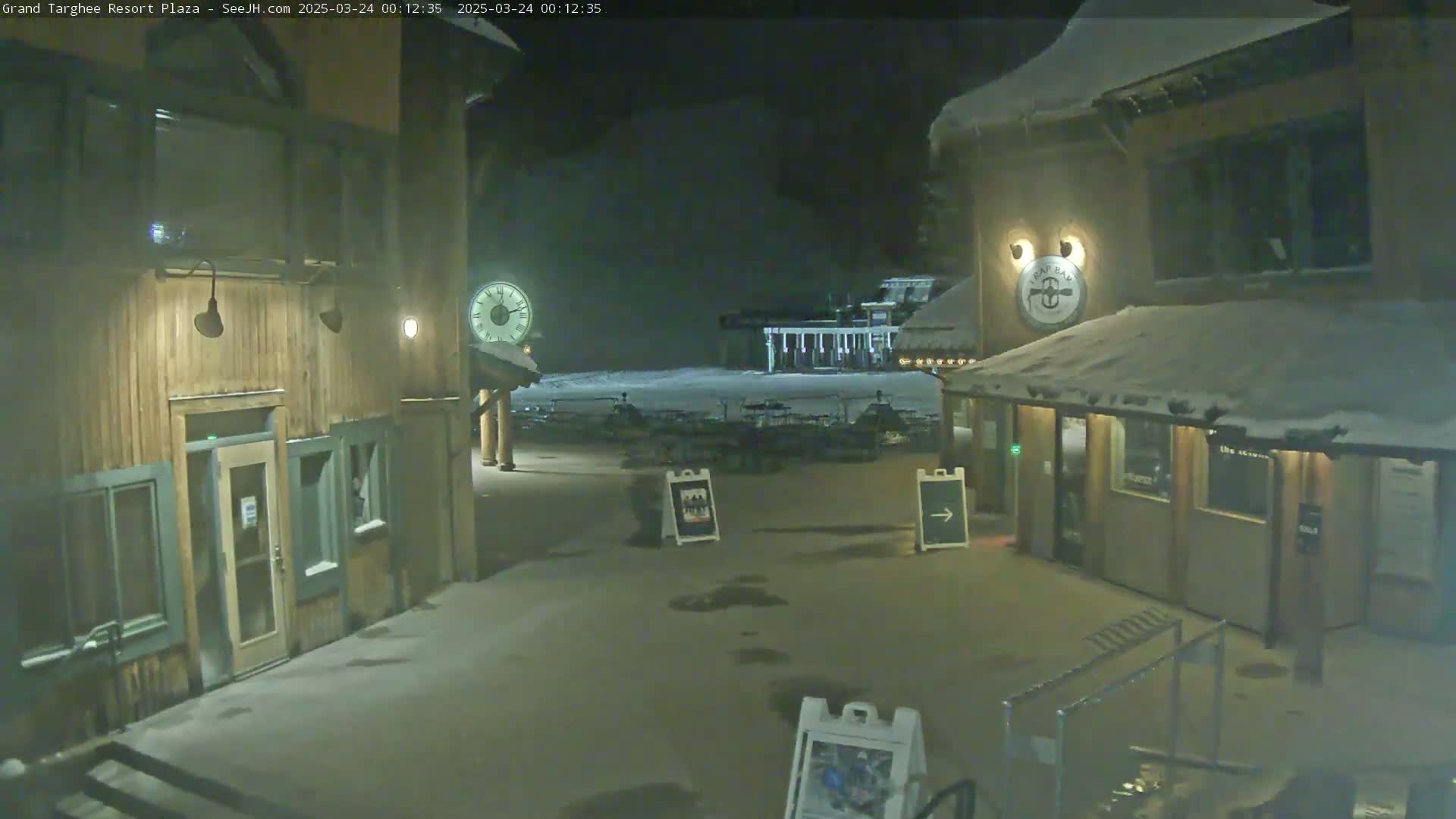 A snow-covered plaza at night, with wooden buildings and a clock on one side, is illuminated by lights, with a distant structure faintly visible in the background.