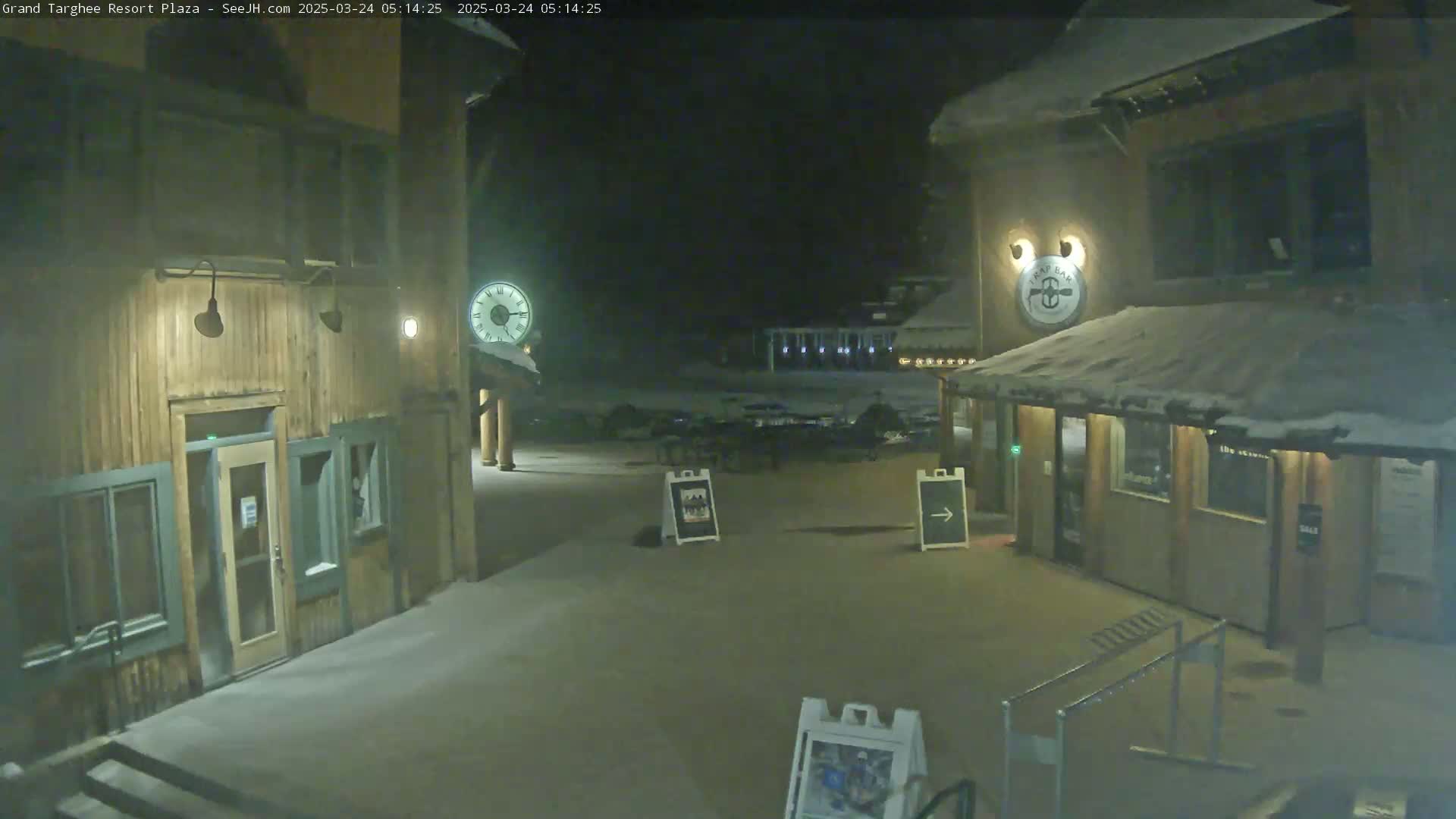 A snow-covered plaza at night, illuminated by lights on wooden buildings, shows a clock and several signs.
