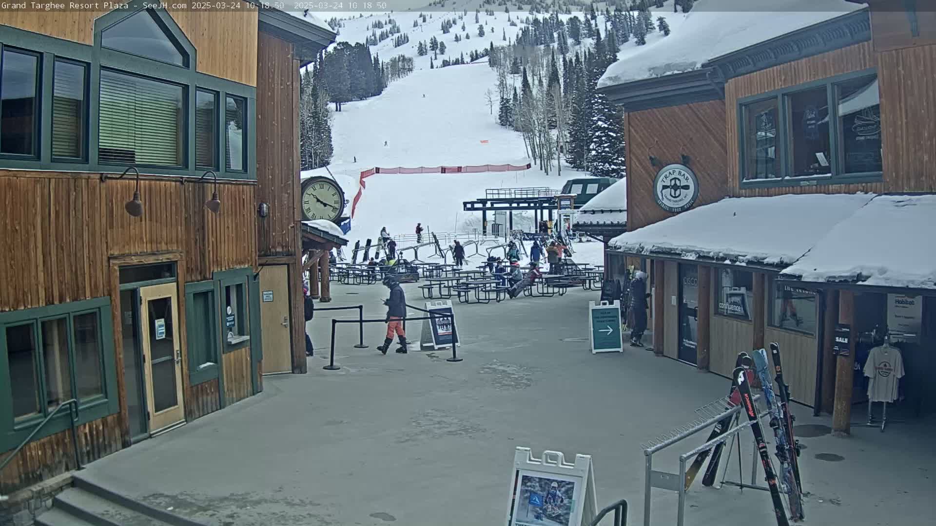 A plaza at a ski resort on a snowy day shows several wooden buildings, outdoor seating, a ski lift in the background, and people walking around.