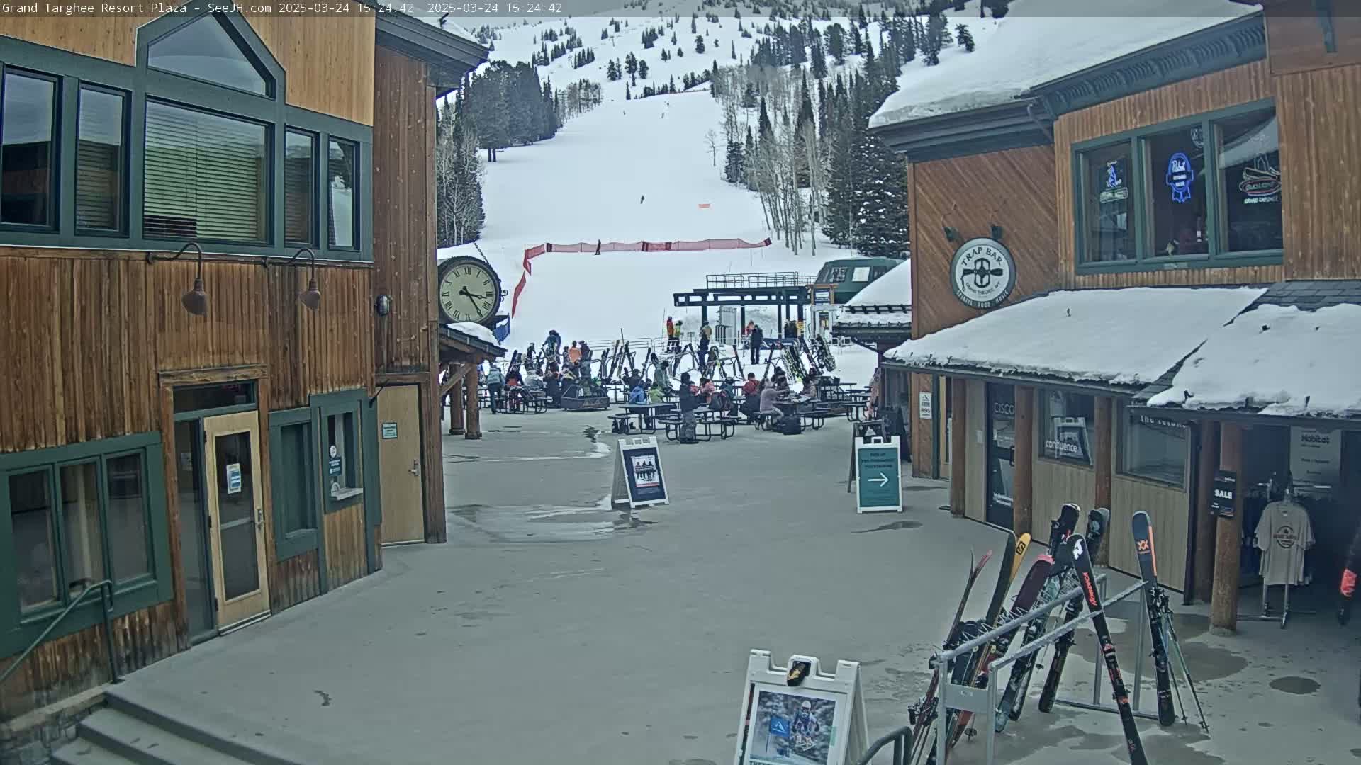 A plaza with wooden buildings, a ski slope in the background, and people sitting at tables under a partly cloudy sky.