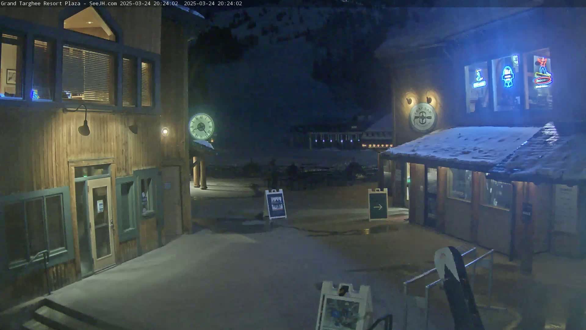 A snow-covered plaza at night, featuring two wooden buildings with illuminated windows and signage, is dimly lit.