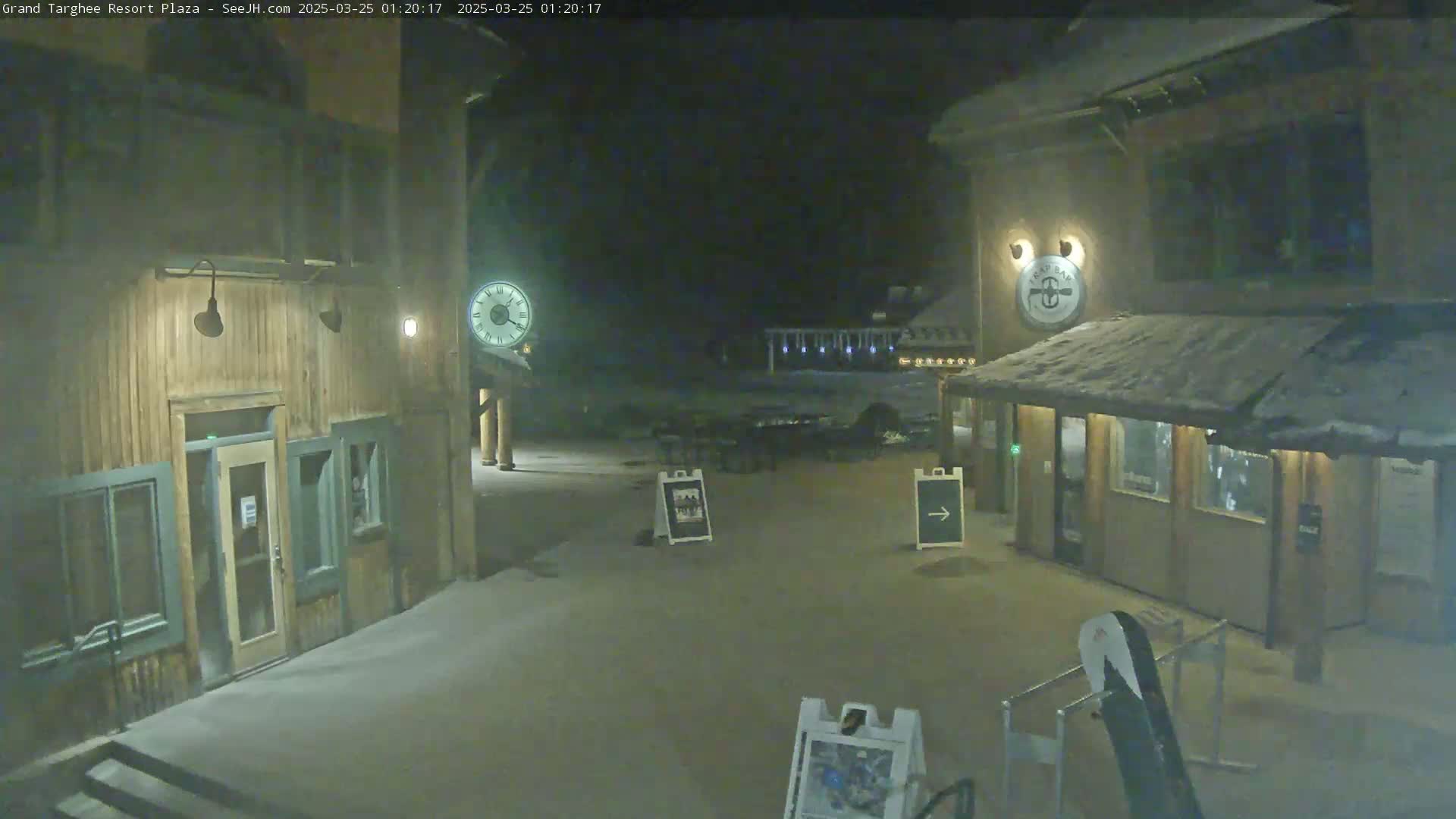 A snow-covered plaza at night, illuminated by dim lights, features two wooden buildings with entrances and signage.