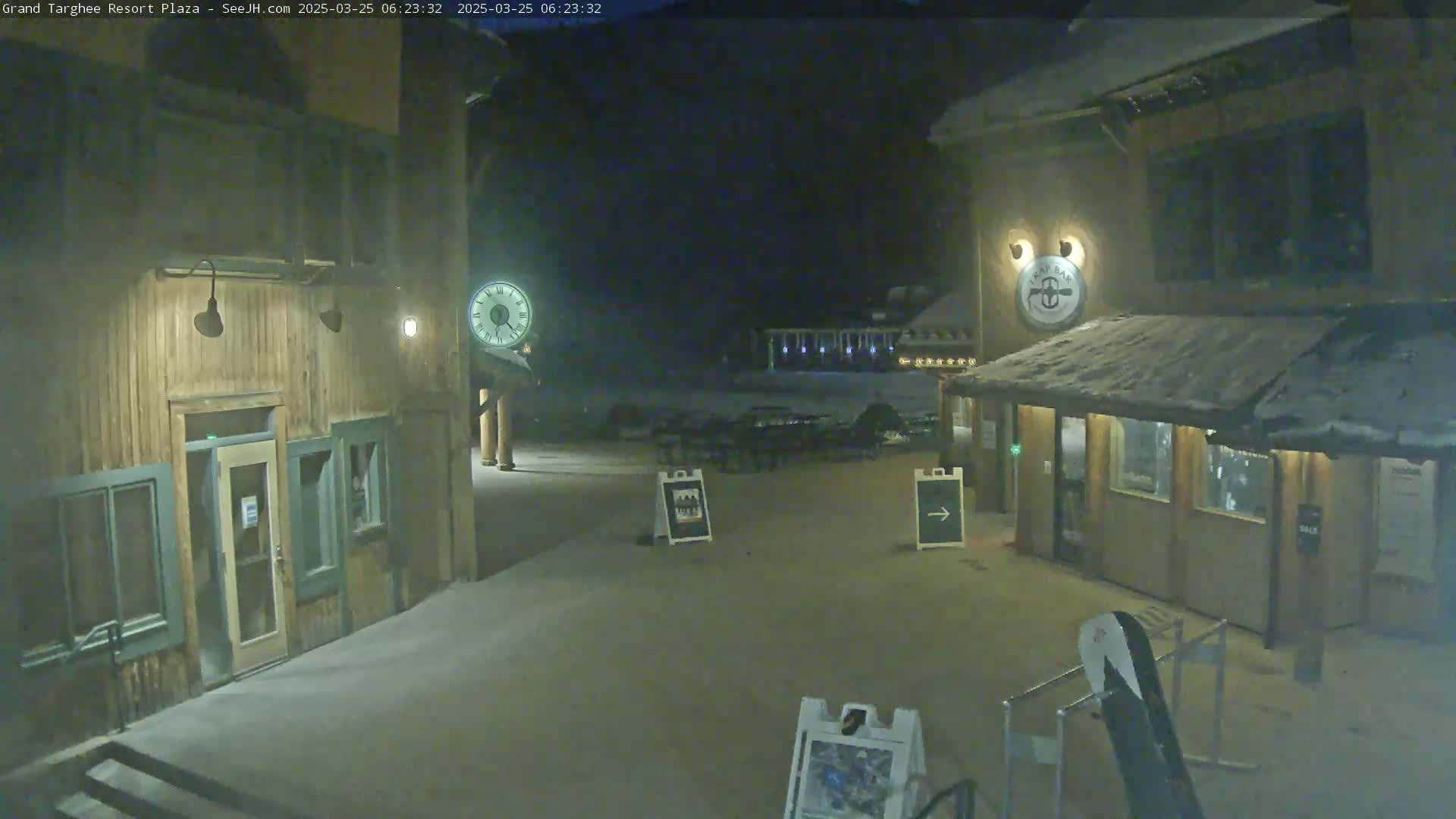 A snow-covered plaza at night, illuminated by lights on wooden buildings, features a clock, signage, and outdoor seating.