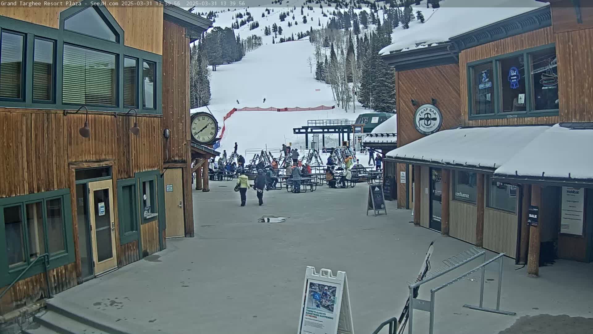 A plaza at a ski resort, with snow on the ground and surrounding buildings, shows several people gathered at tables, while a ski slope is visible in the background.