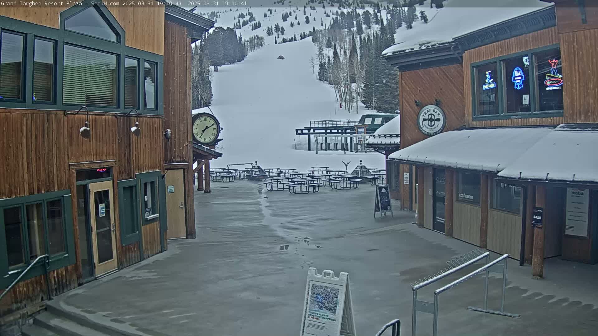 A snow-covered plaza with wooden buildings, outdoor seating, and a ski lift in the background.