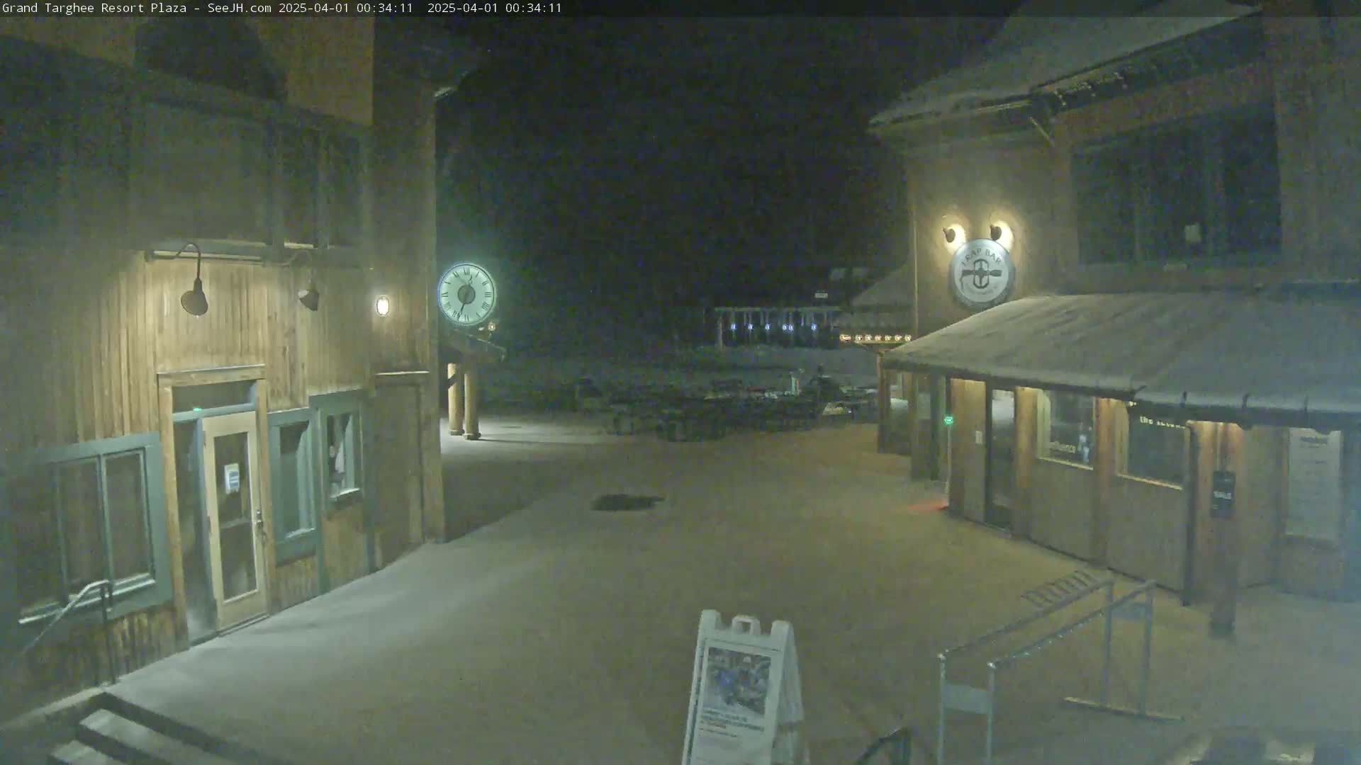 A snow-covered plaza at night is illuminated by lights on wooden buildings, one of which displays a clock.