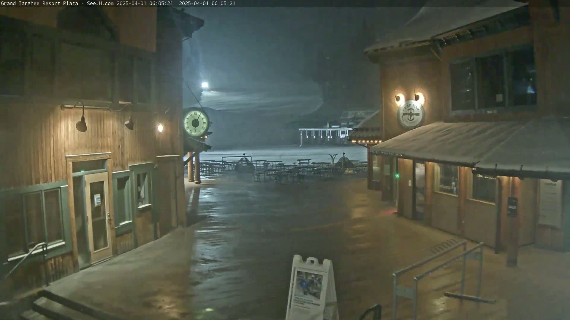 A snow-covered plaza at night, illuminated by lights on wooden buildings, with empty picnic tables visible in the distance.
