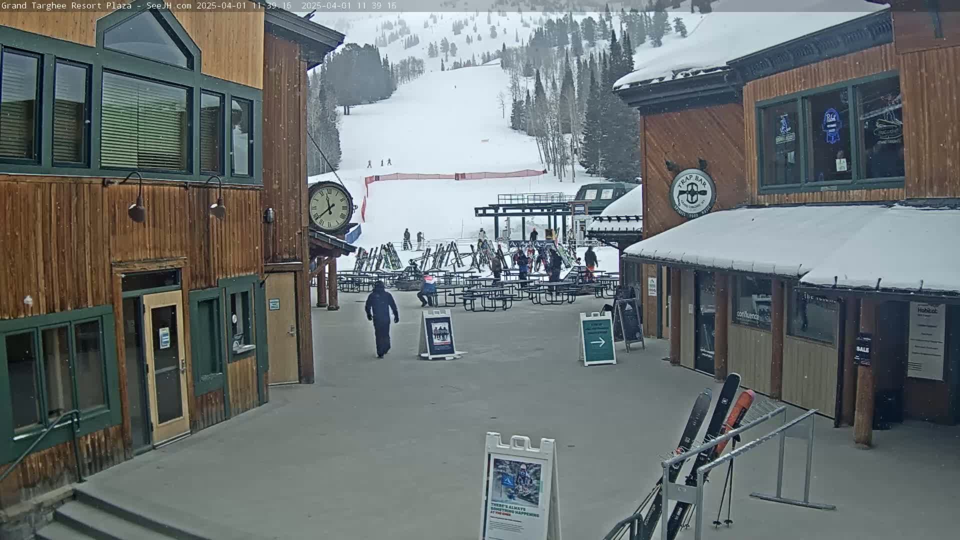 A plaza at a ski resort, lightly snowing, shows wooden buildings, a clock, outdoor seating, and a ski slope in the background.