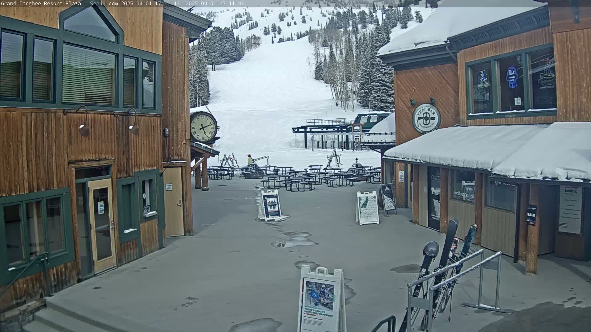 A snow-covered plaza with wooden buildings, outdoor seating, and a ski lift visible in the background under a cloudy sky.