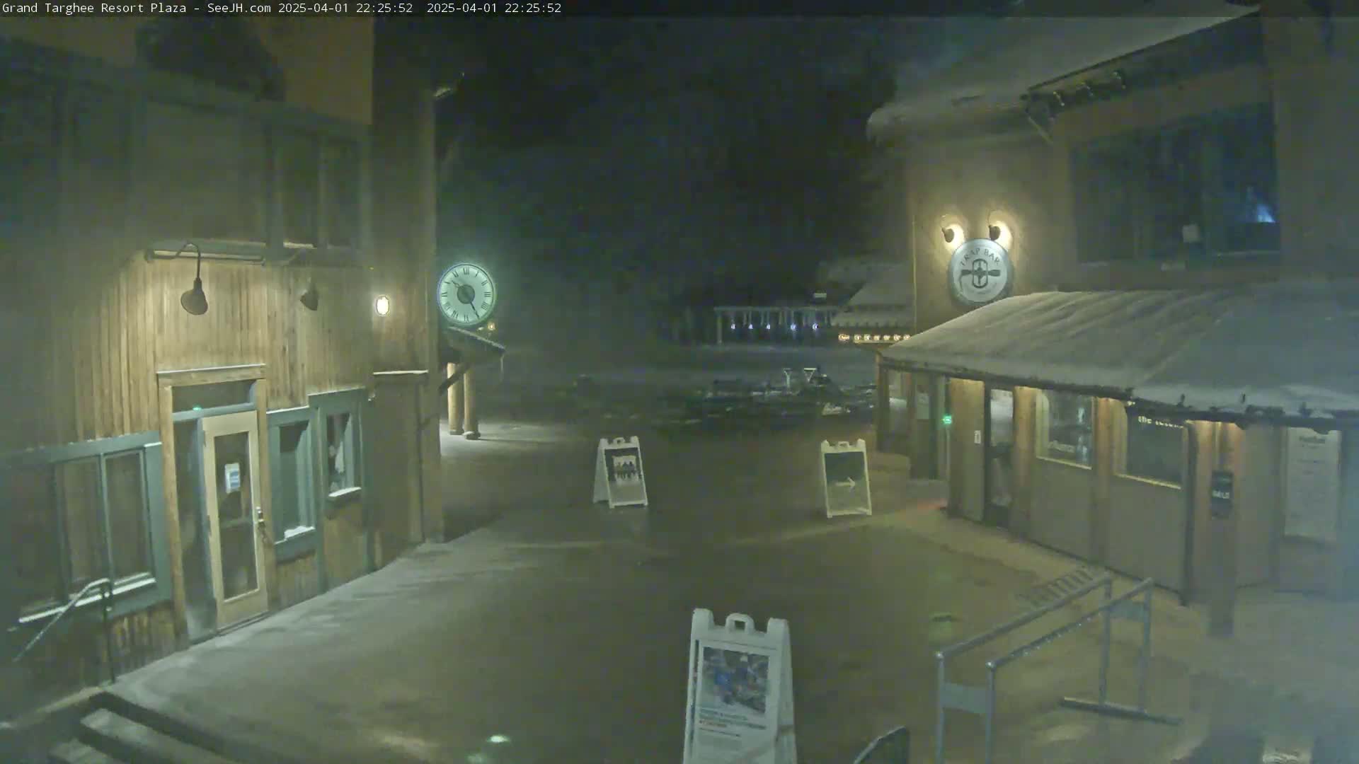 A snow-covered plaza at night is viewed between two wooden buildings, one of which displays a clock.