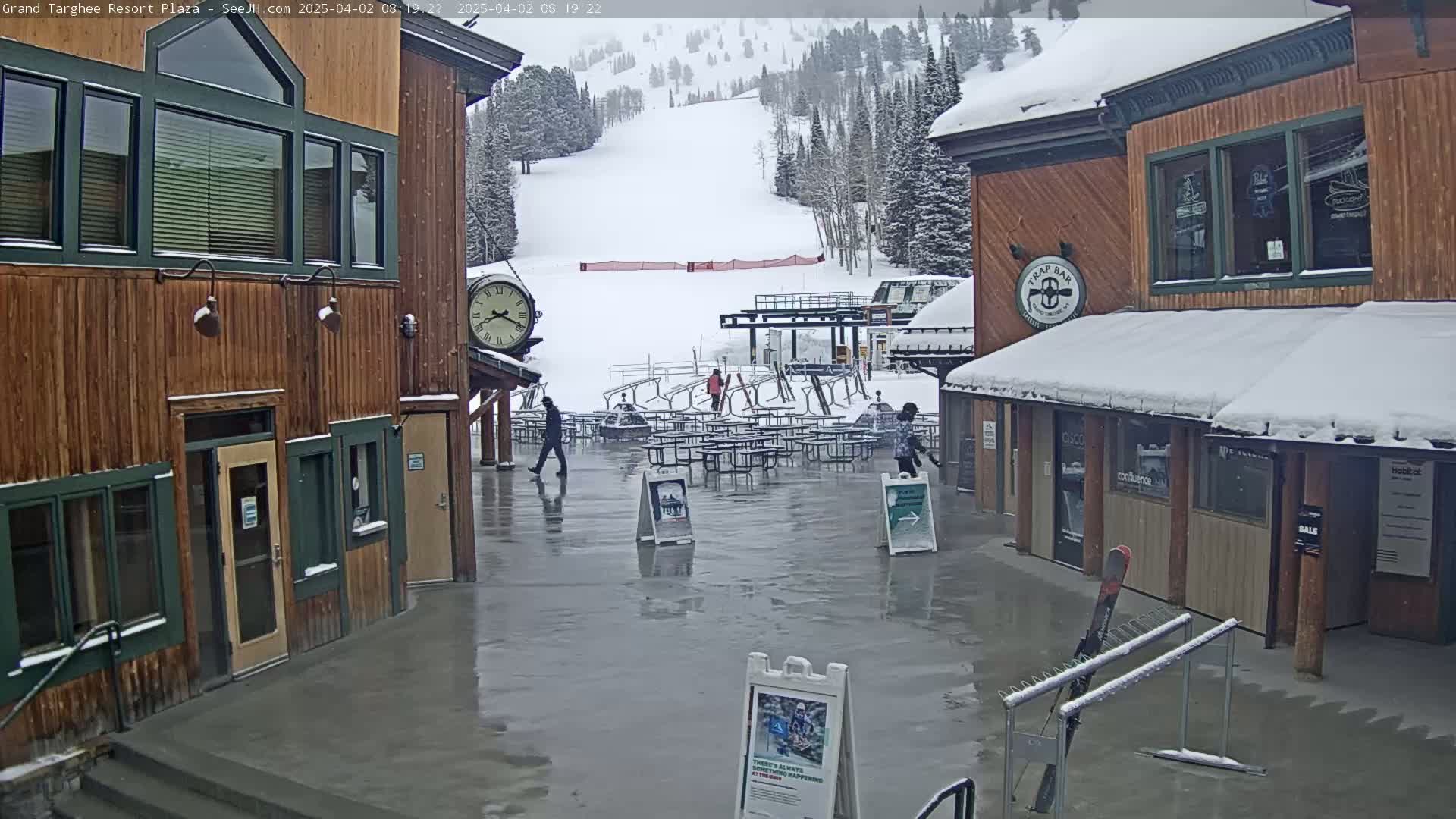 A plaza between two wooden buildings, one with a clock, overlooks a snow-covered ski slope on an overcast day.