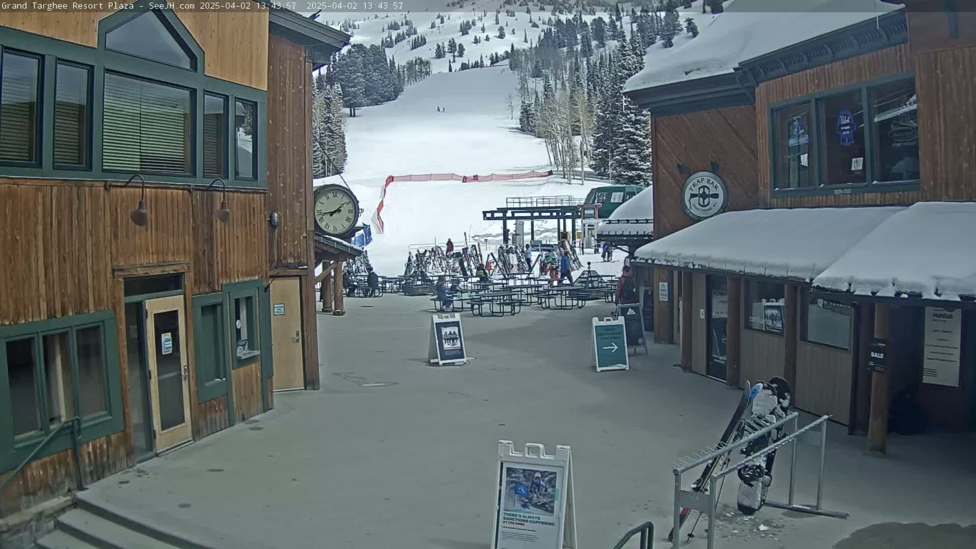 A snow-covered plaza at a ski resort shows two wooden buildings with a clock, outdoor seating, and a ski slope in the background.