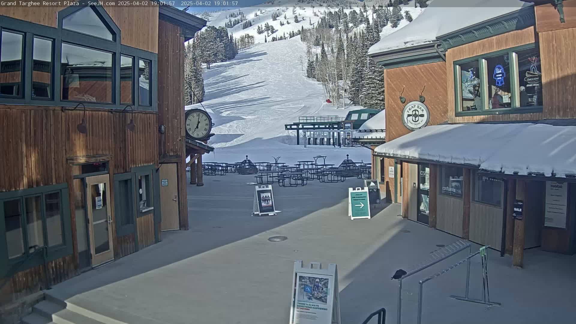A plaza with wooden buildings, snow-covered ground, and a ski slope in the background under sunny conditions.