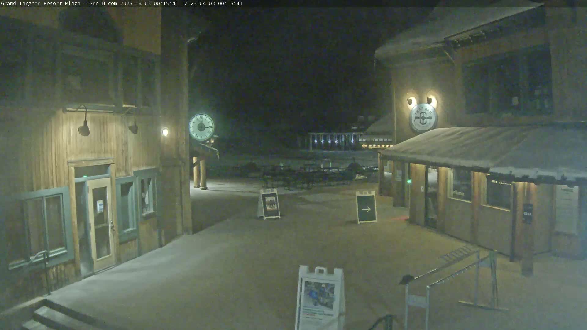 A snow-covered plaza at night, featuring two wooden buildings with lights, a clock, and some outdoor furniture.