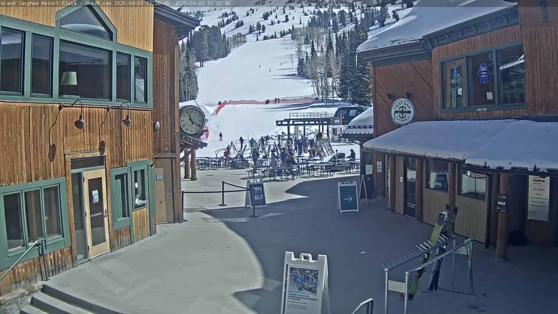 A sunny day at a ski resort plaza shows people sitting at outdoor tables, skiers on the slopes in the background, and wooden buildings with snow on their roofs.