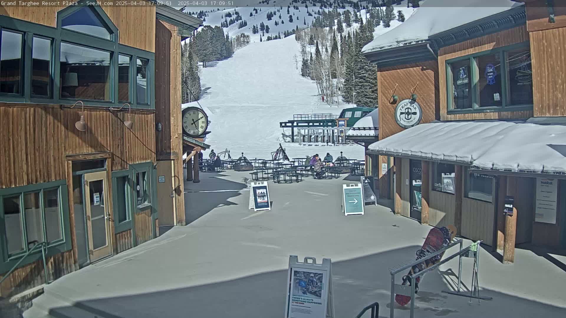A sunny outdoor plaza scene shows several wooden buildings with a ski slope and lift in the background.