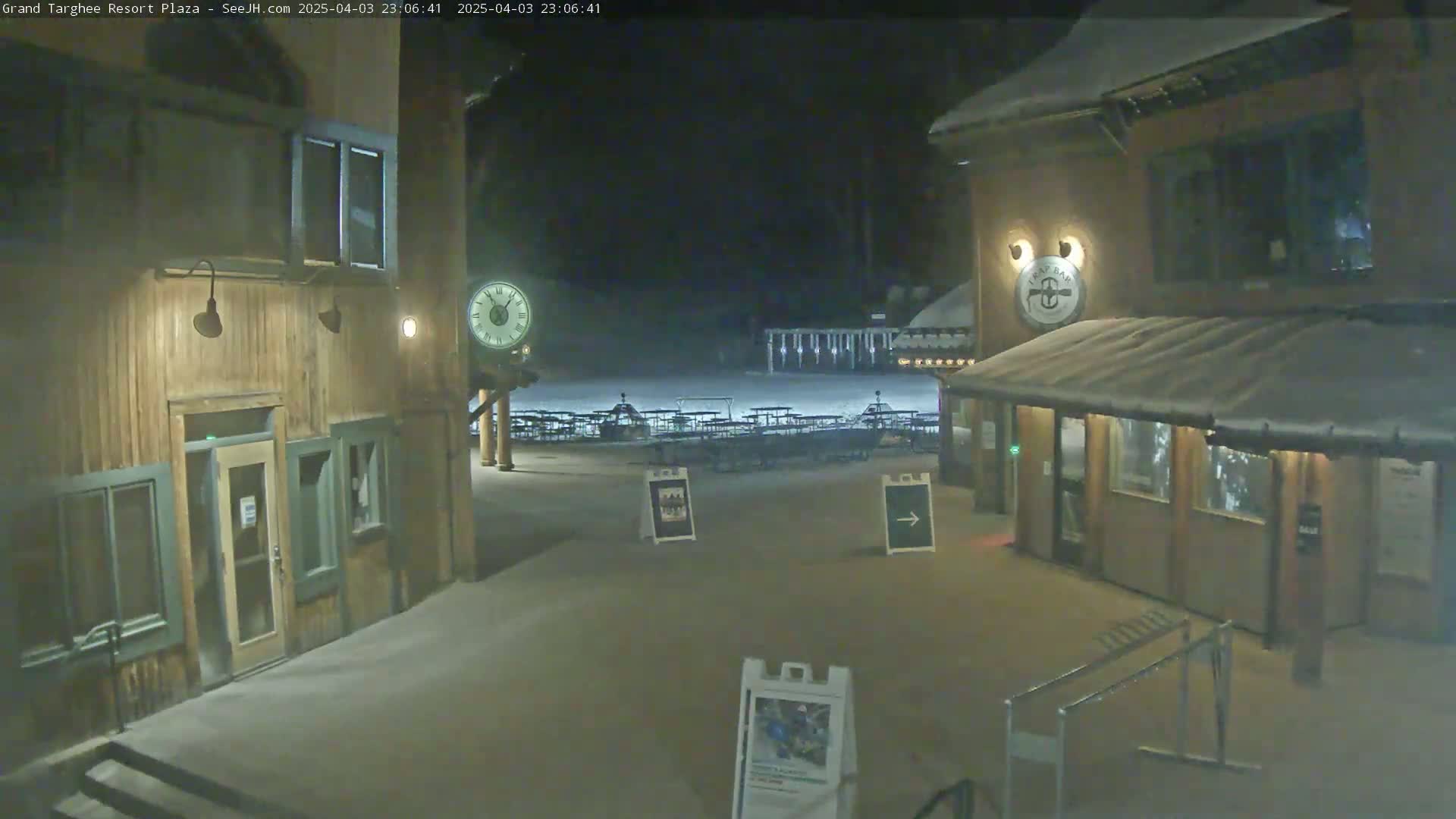A snow-covered plaza at night, illuminated by lights, features wooden buildings with signs, an analog clock, and several empty tables.
