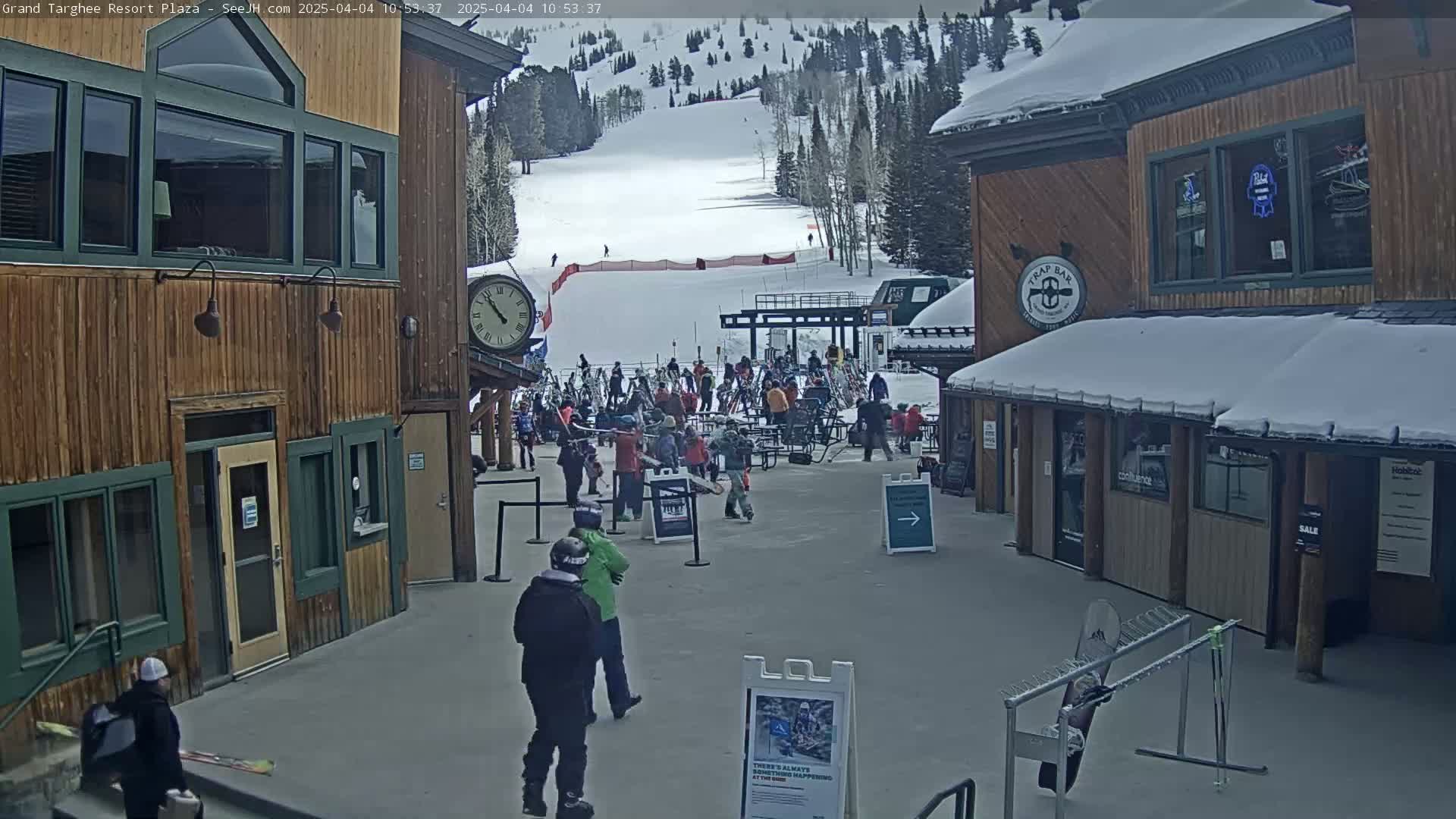 A plaza with several wooden buildings, some people, and a ski slope in the background on a partly cloudy day.