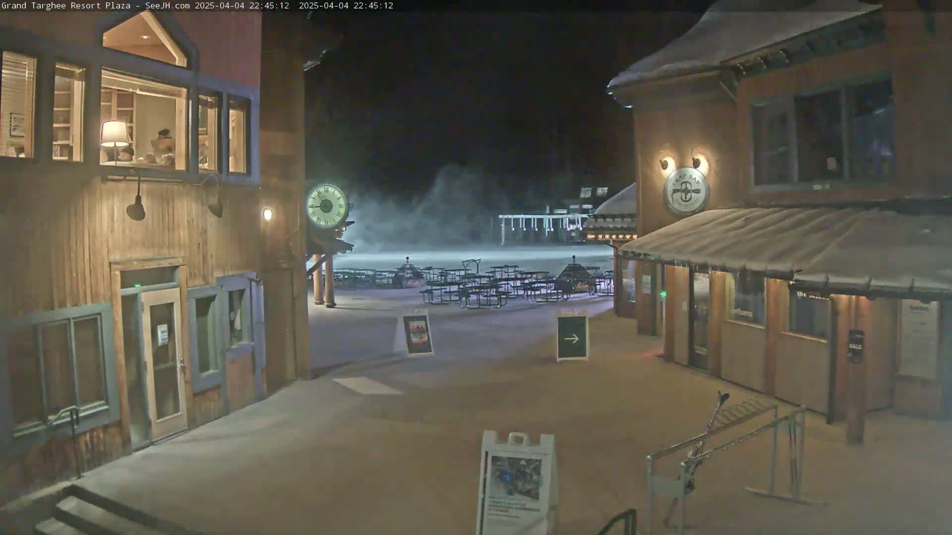 A snow-covered plaza at night, with wooden buildings, empty outdoor seating, and a clock tower.