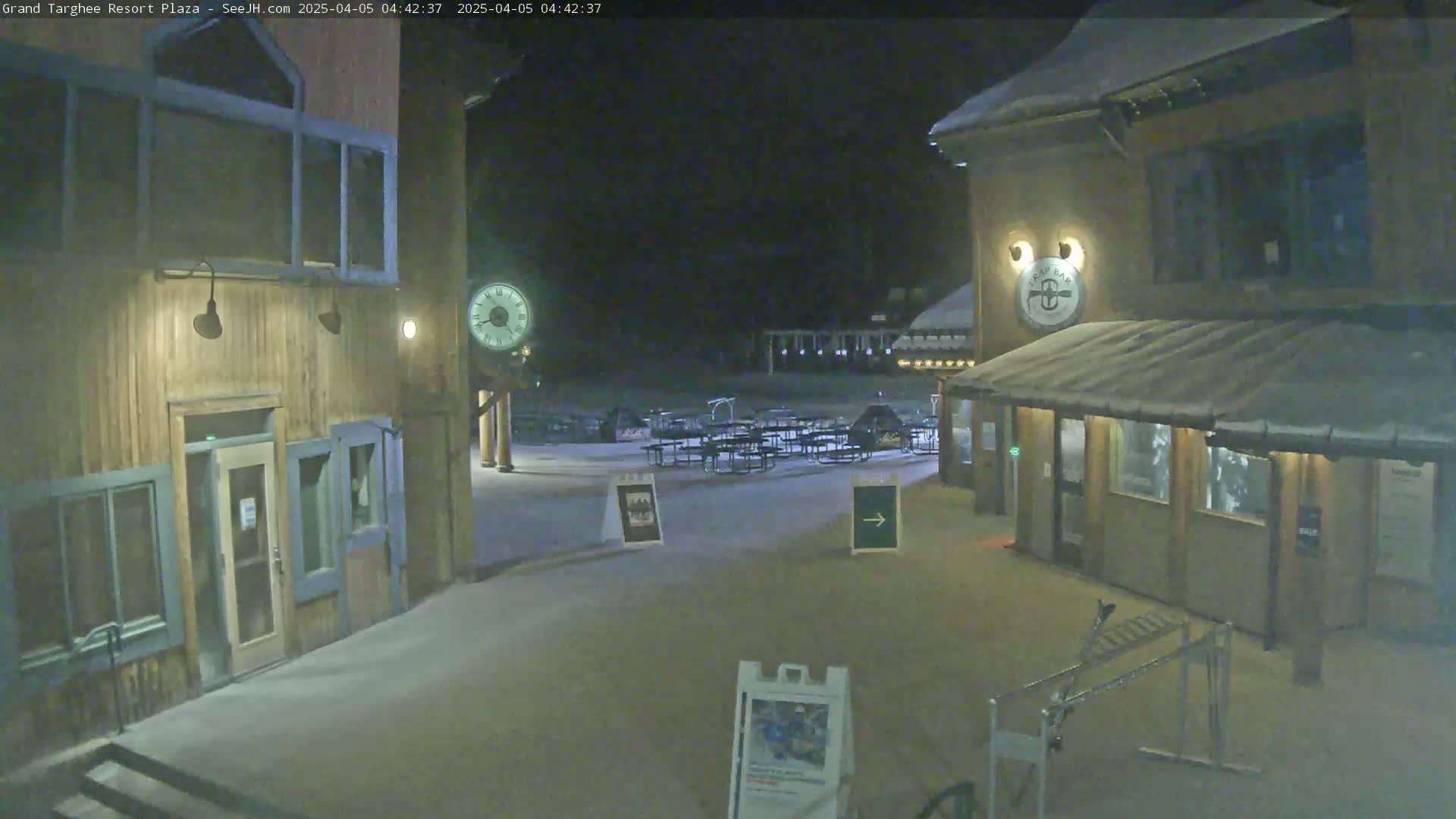 A snow-covered plaza at night, with wooden buildings, outdoor seating, and a clock tower.