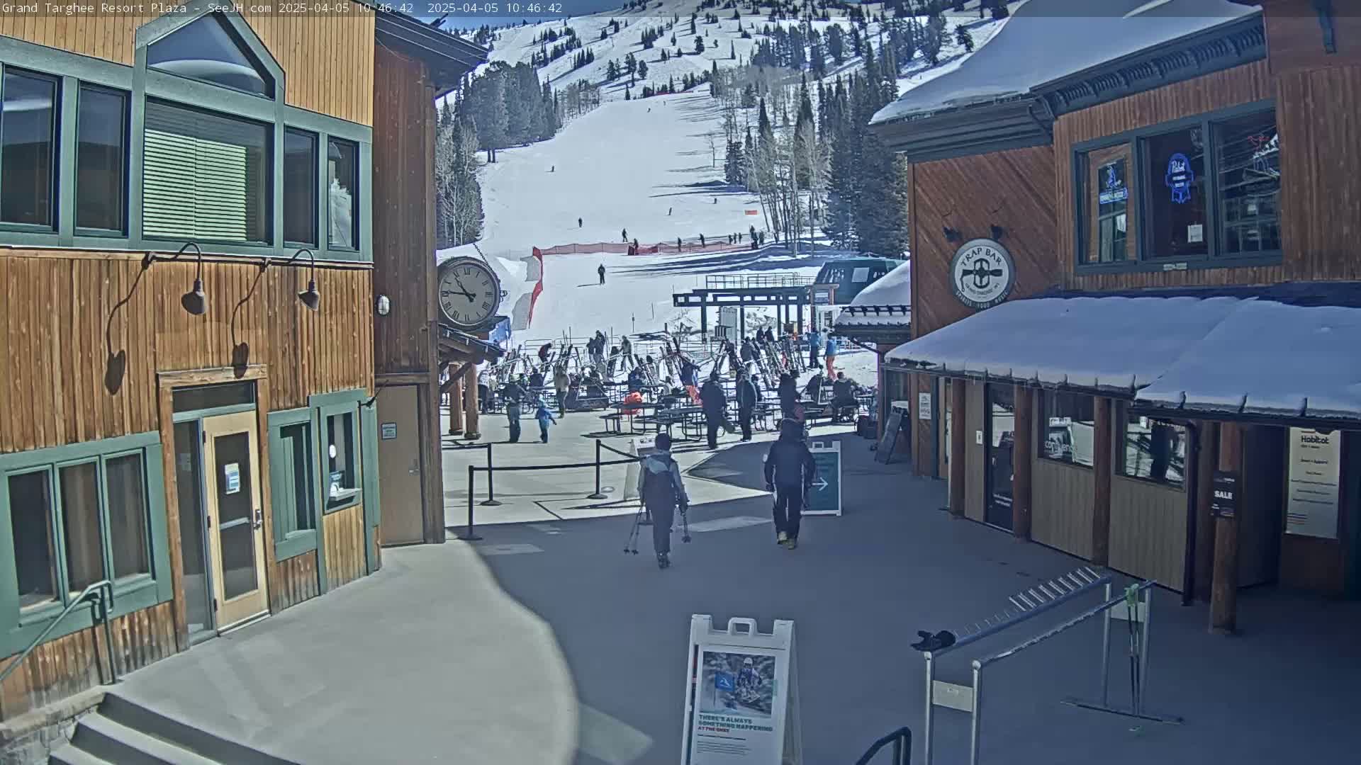 A sunny outdoor plaza at a ski resort shows several people walking near wooden buildings with snow on the roofs and a ski slope in the background.
