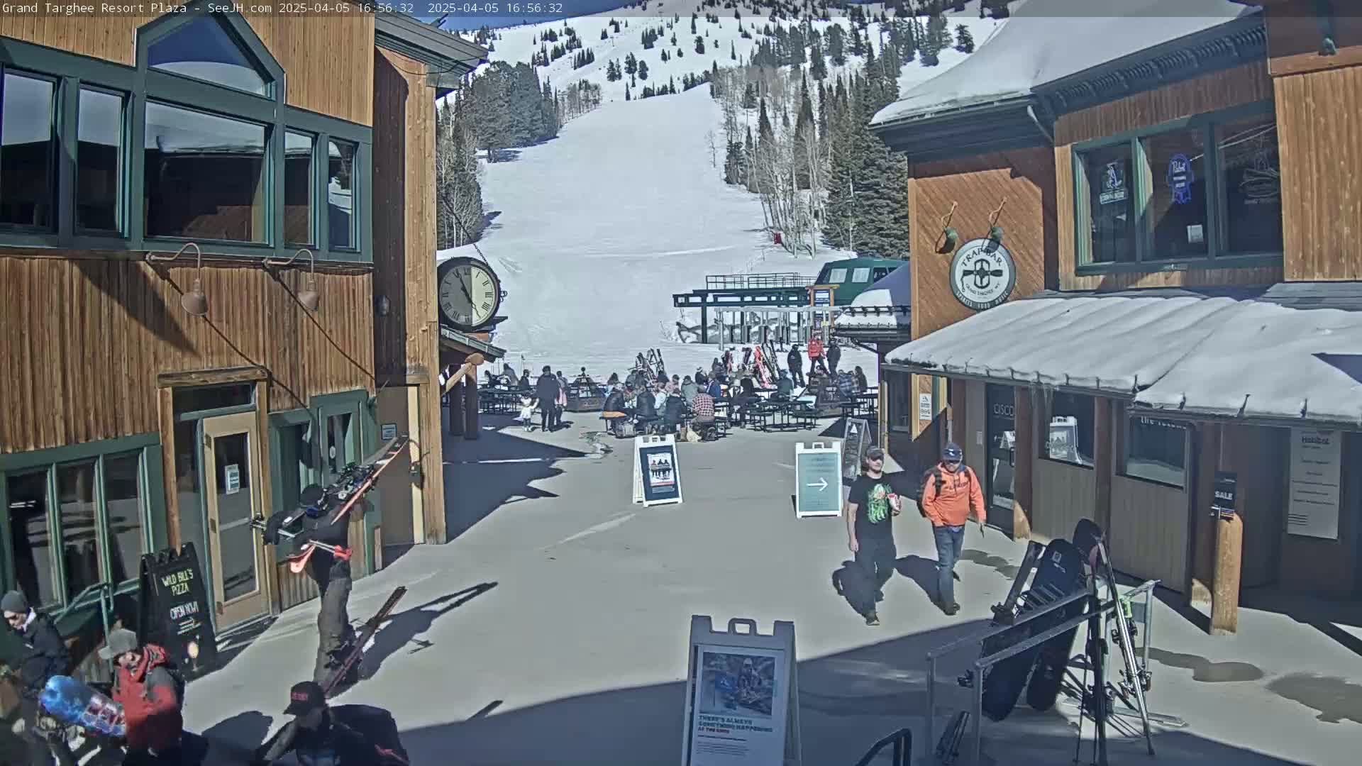 A sunny plaza at a ski resort shows several people milling about near buildings and a ski lift with a snowy mountain in the background.