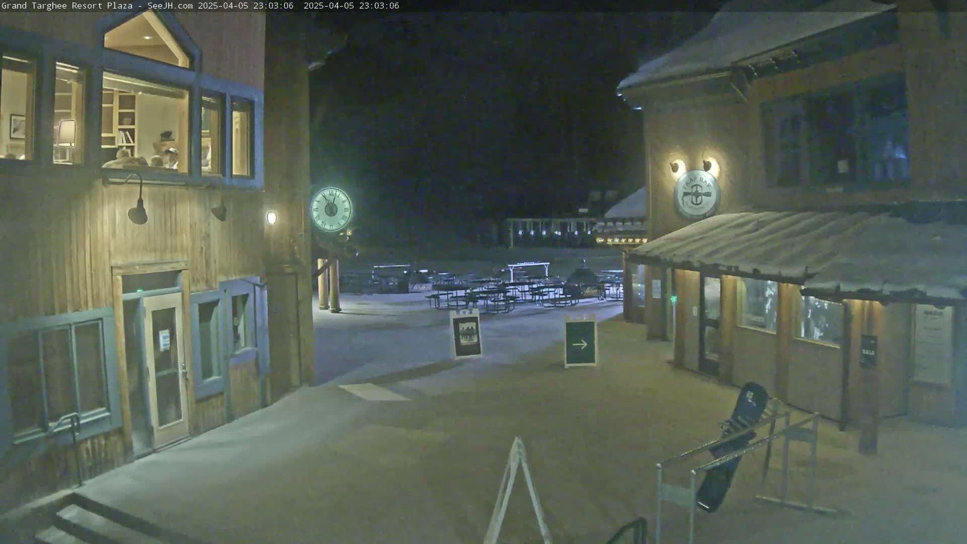 A nighttime view of a snow-covered plaza with wooden buildings, outdoor seating, and a clock.