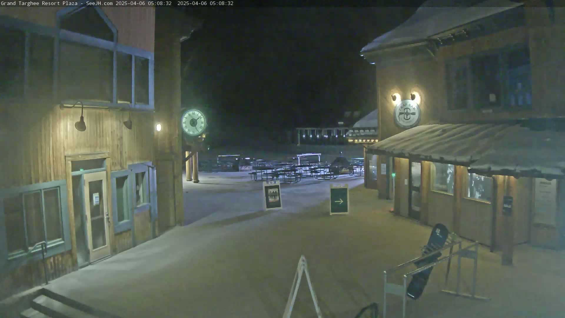 A snow-covered plaza at night, illuminated by lights, shows several wooden buildings with a clock on one, picnic tables, and a snowboard leaning against a rack.