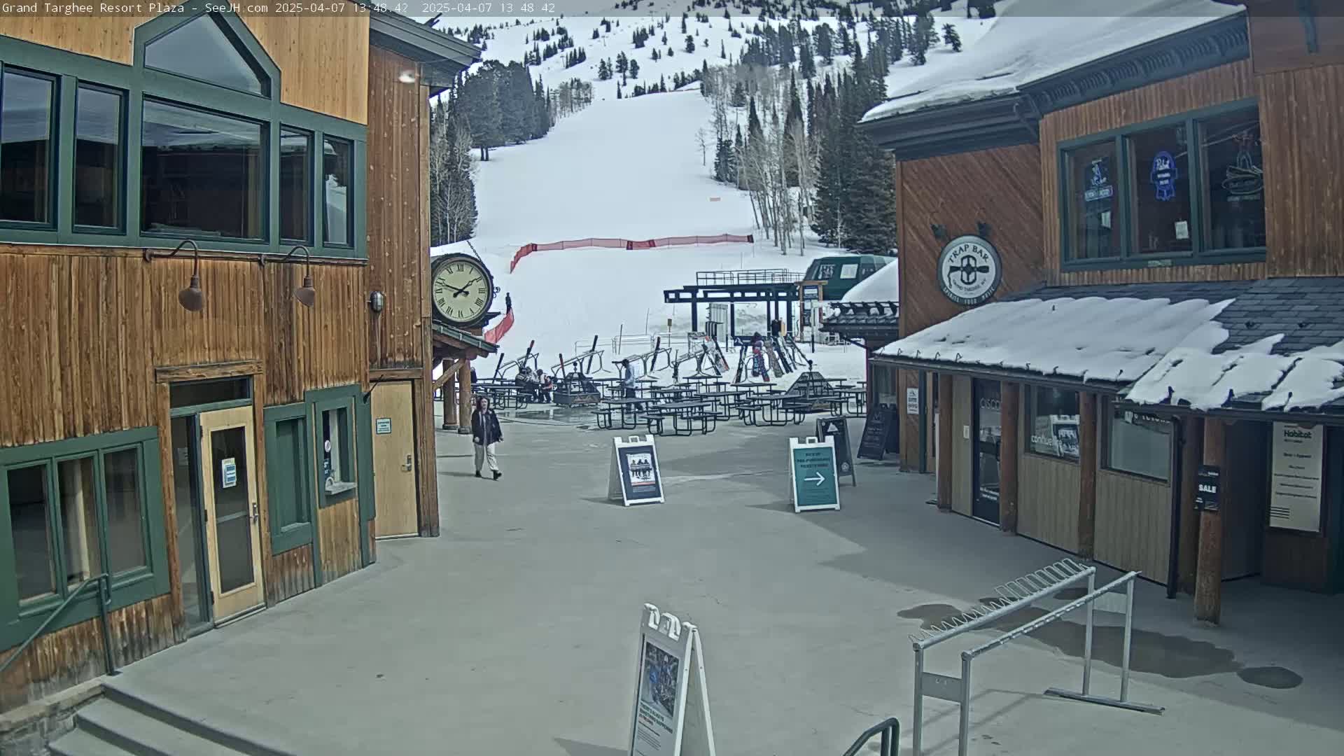 A plaza with wooden buildings, an outdoor clock, several picnic tables, and a ski slope visible in the background under a partly cloudy sky.