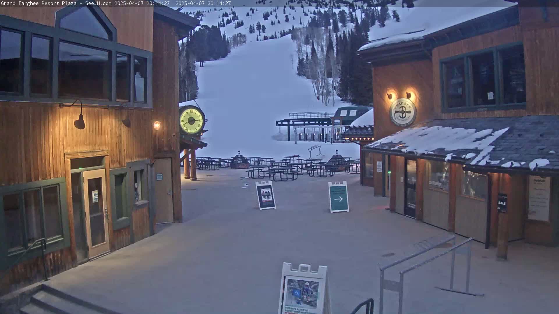 A snow-covered plaza at dusk features several wooden buildings, picnic tables, and a ski lift in the background under a twilight sky.
