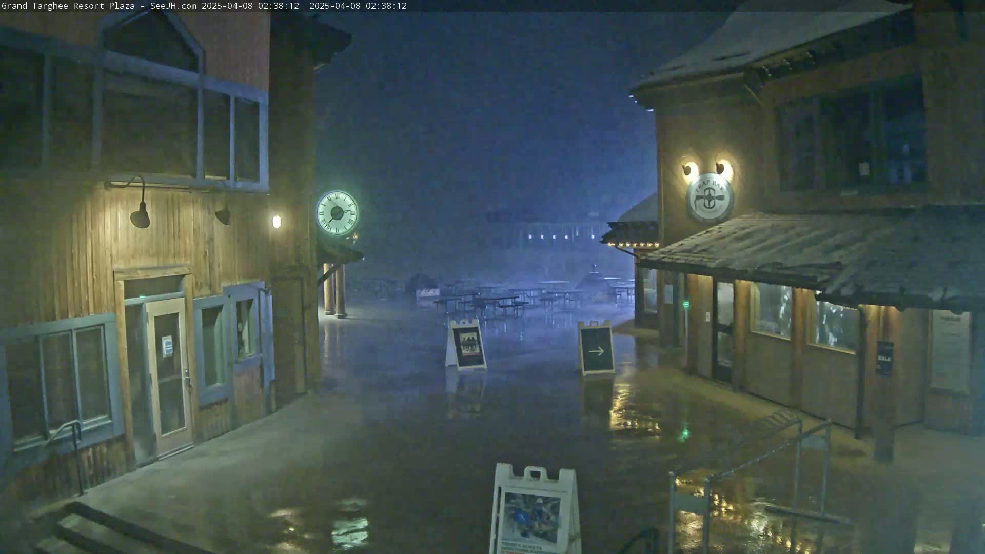 A plaza with wooden buildings and outdoor seating is flooded under a dark, snowy night.