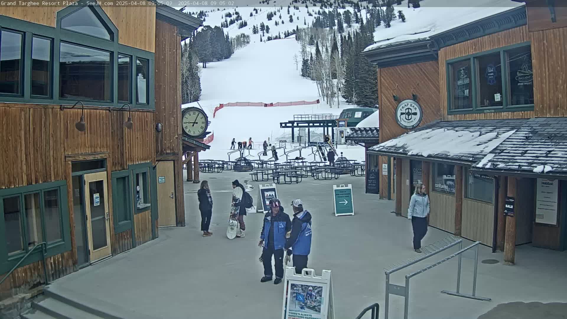 A plaza with wooden buildings, a ski slope in the background, and several people milling about on a snowy day.
