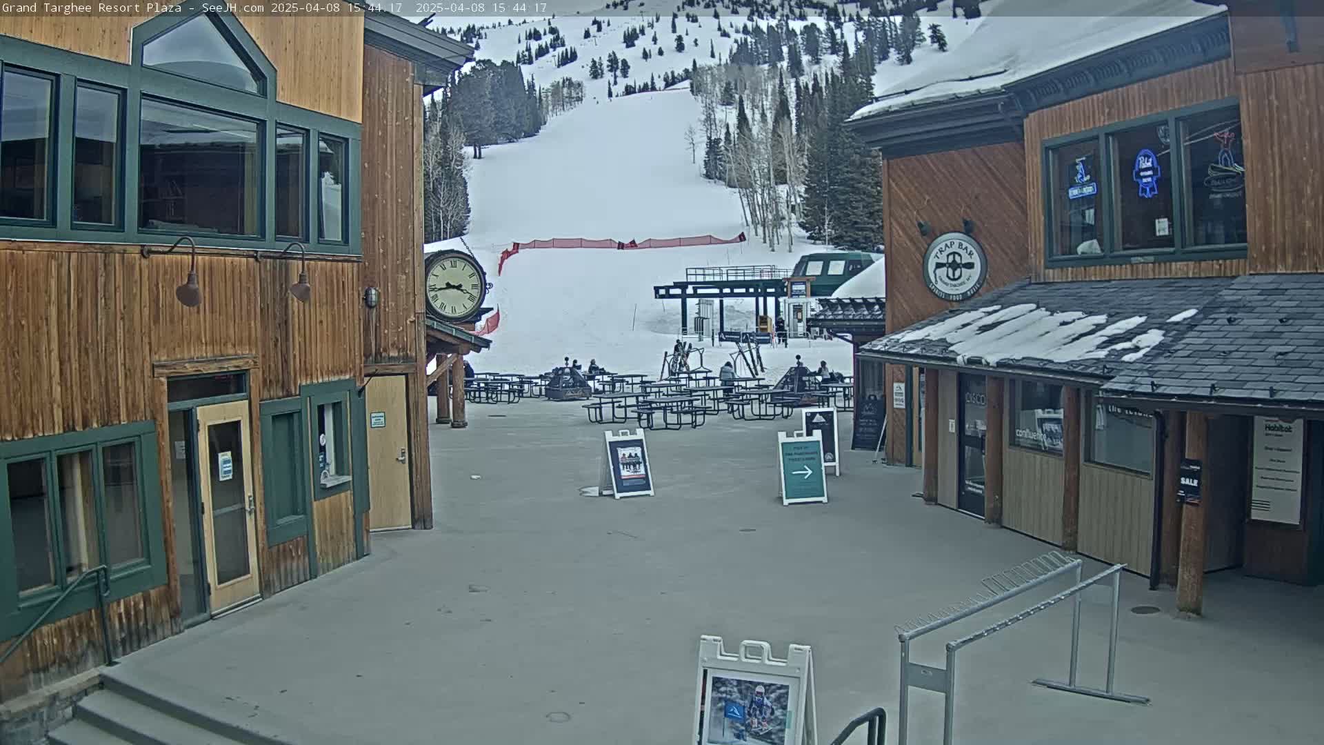 A plaza with wooden buildings, picnic tables, and a ski lift in the background on a partly cloudy day with snow on the ground and roofs.