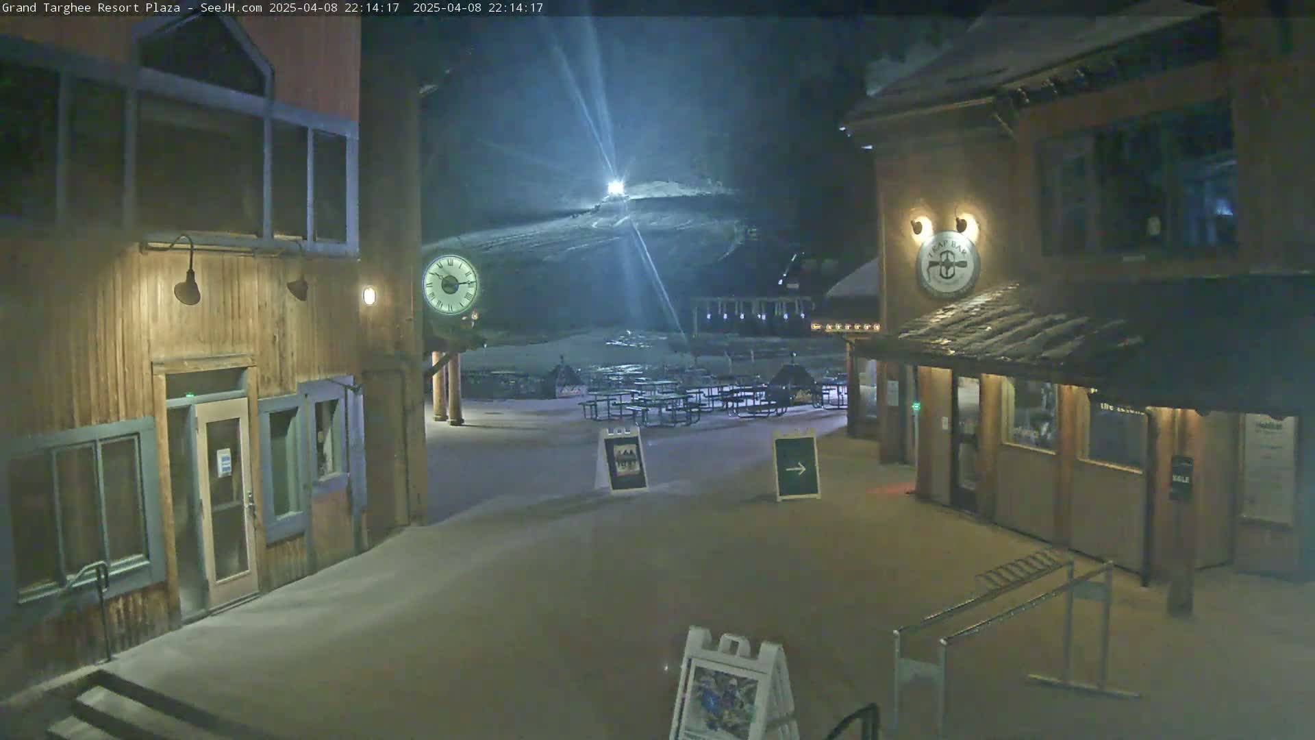 A snow-covered plaza at night, illuminated by lights, shows wooden buildings with some outdoor seating and a hill in the background with lights on it.